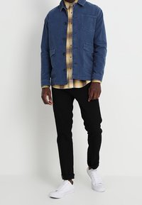 Giacca di cotone blu con tasche frontali, abbinata a una camicia a quadri gialla, pantaloni neri e sneakers bianche. Il modello è in piedi contro uno sfondo semplice.