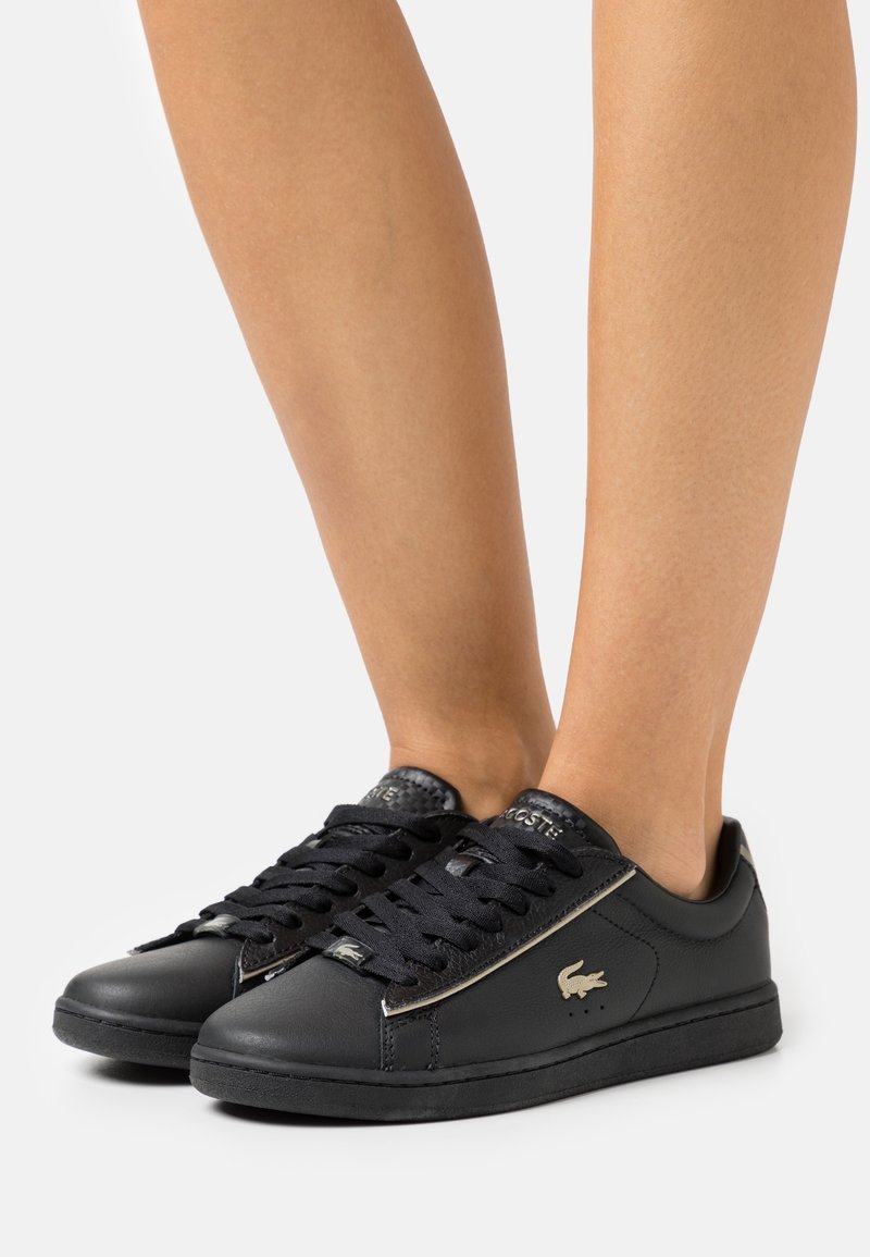 Baskets basses noires Lacoste avec des lacets noirs portées pieds nus, montrant le logo crocodile doré sur le côté et la languette.