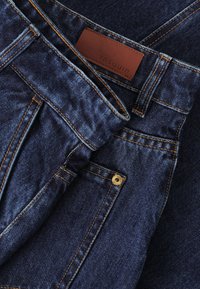 Jeans de mezclilla azul oscuro con costuras naranjas, que presentan una etiqueta de cuero etiquetada "TNTUUM" y un remache metálico dorado en el bolsillo.