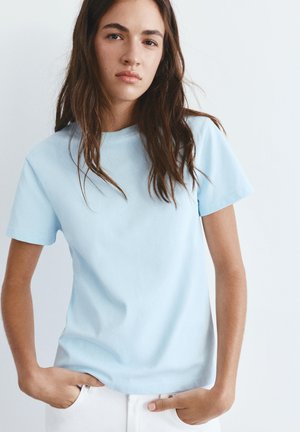 Mladá žena s dlouhými hnědými vlasy, oblečená v světle modrém tričku s krátkým rukávem a bílých kalhotách, s rukama v kapsách, na světlém jednolitějším pozadí.