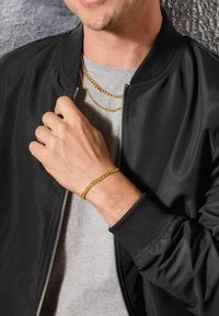 Collana e bracciale in oro indossati da una persona con una maglietta grigia e una giacca bomber nera, che mostrano texture morbide e finiture lucide.