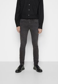 Hombre vistiendo jeans oscuros de corte ajustado, camisa negra de botones y zapatos de cuero negros con cordones, de pie contra un fondo blanco.