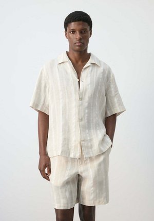 Man wearing beige short-sleeve button-up shirt and matching shorts with vertical stripes, standing against a plain white background.