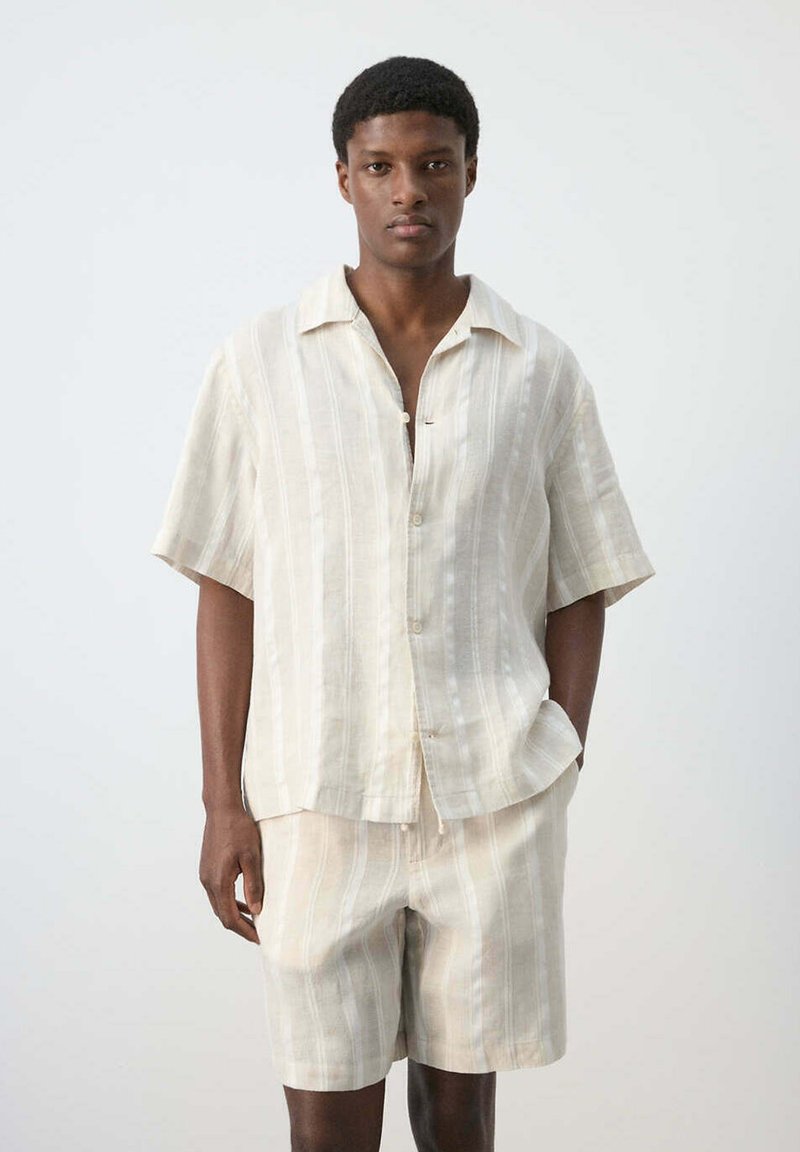 Homme portant une chemise beige à manches courtes boutonnée et un short assorti à rayures verticales, debout devant un fond blanc uni.