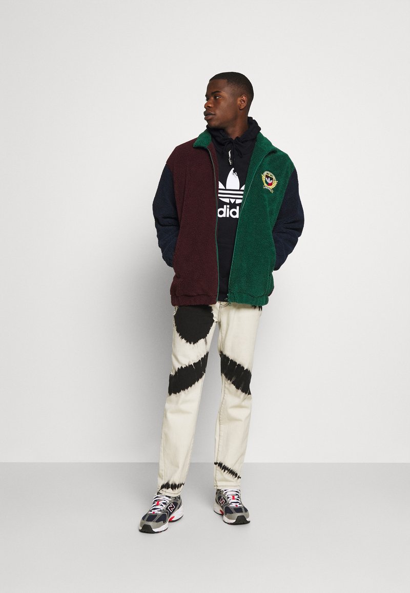 Fleece striukė su vyno raudonais ir žaliais šonais, derinama su juoda džemperiu su gaubtu, raštuotomis kelnėmis ir spalvingais sportbačiais, demonstravusi tekstūrinius audinius.