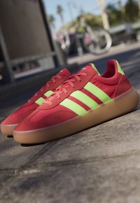 Red and green Adidas sneakers with translucent gum soles placed on outdoor pavement in daylight.