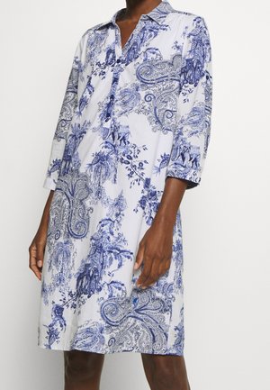 Witte jurk tot de knie met blauw gedetailleerde bloemen- en olifantenpatronen, driekwartmouwen en een knoopsluiting, gedragen door een persoon met een donkere huidskleur.