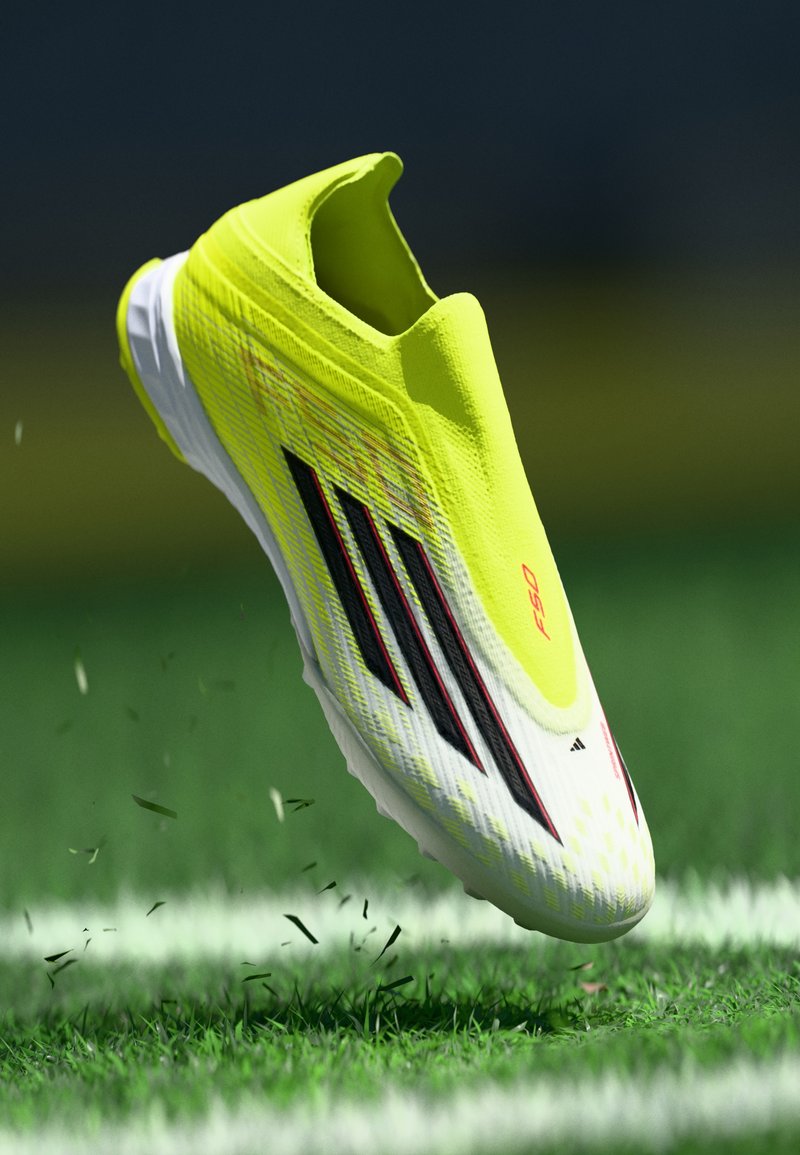 Geltonos futbolo bateliai su stilingu dizainu, lengva medžiaga, tekstūruota viršūne, balta padais ir juodais juostelėmis. Padėti virš žolės.