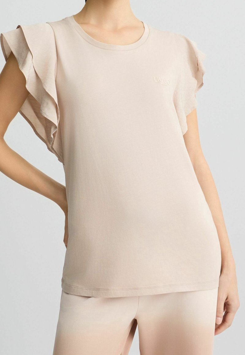 Femme portant un haut beige à manches courtes avec des manches à volants superposés et un pantalon beige clair assorti, main posée sur la hanche.