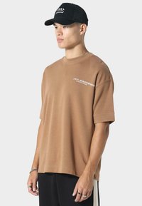Ung mand iført en brun oversized T-shirt med hvid tekst og en sort kasket med hvid skrift, stående mod en ensfarvet baggrund.
