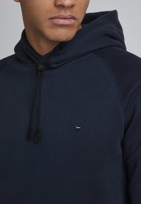 Tamsiai mėlynas džemperis su kapuce, styginiu uždarymu, šoninėmis detalėmis ir mažu trikampio formos logotipu ant krūtinės. Minkšta, tekstūrinė medžiaga.