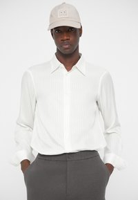 Hvitstripet langermet skjorte med spiss krage, sammen med grå bukser og en beige caps med et logo-patch.