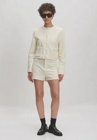 Crèmekleurige tweedelige outfit met een cropped jack met knoopsluitingen en twee voorzakken, gecombineerd met bijpassende shorts en loafers.