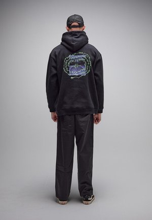 Osoba ubrana w czarną bluzę z kapturem z kolorowym nadrukiem i wzorem na plecach, czarne spodnie, czarna czapka i czarno-białe trampki, stojąca tyłem.