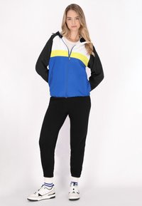 Jachetă windbreaker în culorile albastru, alb, galben și negru; cu fermoar în față, glugă și manșete ribbed, asortată cu pantaloni jogging negri și adidași albi.