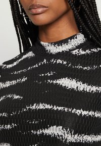 Gros plan sur une personne portant un haut à col montant texturé noir et blanc avec des cheveux tressés et des boucles d'oreilles créoles.