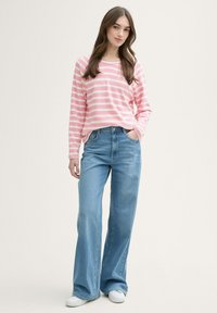 Camisa de rayas rosa y blanca de manga larga combinada con vaqueros acampanados de color azul claro. Modelo luciendo zapatillas blancas, de pie con una mano en el bolsillo.