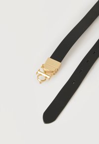 Μαύρη δερμάτινη ζώνη με χρυσή μεταλλική αγκράφα που διαθέτει σχέδιο με πλεγμένα γράμματα, τοποθετημένη επίπεδα σε λευκή επιφάνεια.