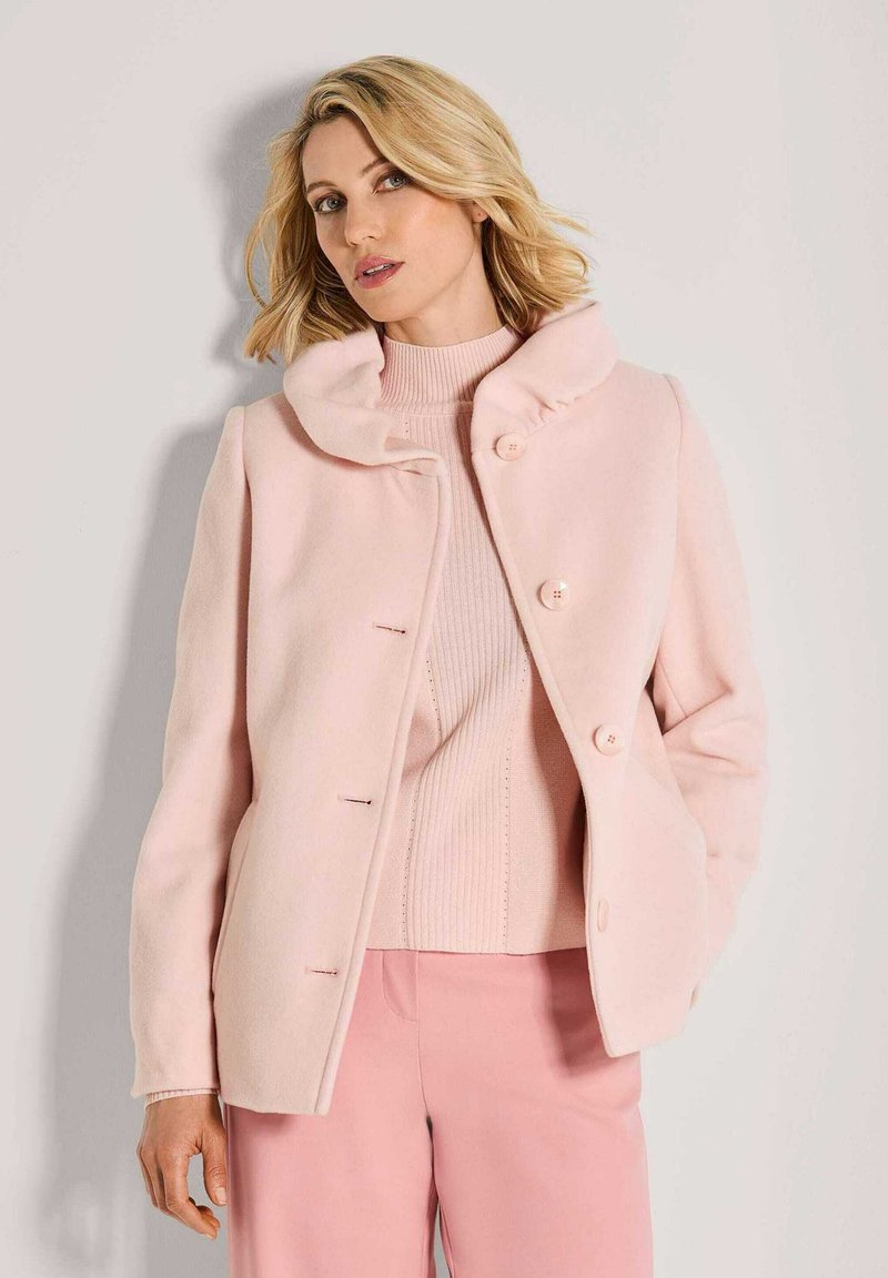 Vrouw in een lichtroze wollen jas over een geribde lichtroze coltrui en bijpassende rose broek, staand tegen een eenvoudige muur.