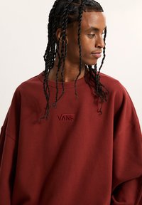 Joven con trenzas que lleva una sudadera ancha de Vans color burdeos, mirando hacia un lado contra un fondo simple.