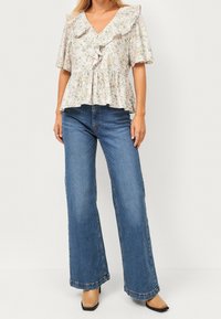 Frau trägt eine blumige Rüschbluse, weite blaue Jeans und beige Schuhe mit quadratischer Zehenform, steht vor einem hellen einfarbigen Hintergrund.