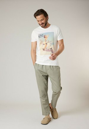 Homme souriant, portant un t-shirt blanc avec un imprimé de scooter vintage, un pantalon gris clair et des chaussures beige sans lacets, debout devant un fond uni.