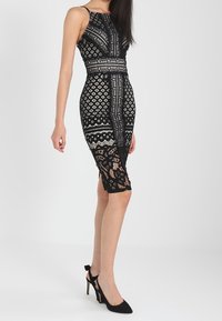 Robe en dentelle noire présentant des motifs géométriques, une silhouette ajustée et des accents transparents à l'ourlet. Assortie avec des chaussures à talons noirs.