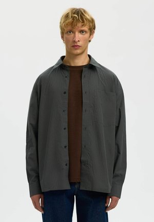 Jeune personne aux cheveux blonds courts portant une chemise gris foncé à boutons, déboutonnée sur un t-shirt marron et un jean bleu, debout sur fond blanc.