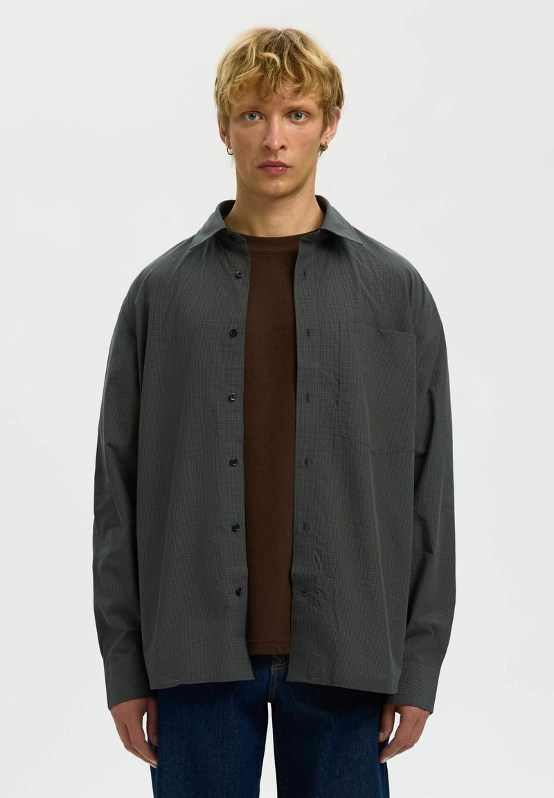 Jeune personne aux cheveux blonds courts portant une chemise gris foncé à boutons, déboutonnée sur un t-shirt marron et un jean bleu, debout sur fond blanc.