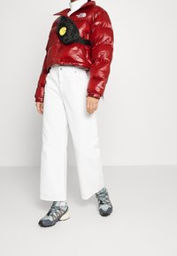 Czerwona kurtka puchowa z wysokim kołnierzem; białe szerokie dżinsy; czarna torebka na ramię z żółtym akcentem; szare i niebieskie buty sportowe z teksturowanymi podeszwami.