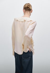 Blusa beige chiaro con colletto arricciato e dettagli a goccia sul retro, abbinata a pantaloni in denim scuro. Tessuto liscio e vestibilità ampia.