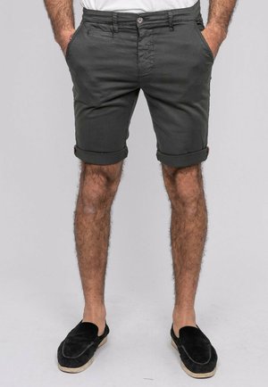Homme debout, les mains dans les poches, portant un short gris foncé retroussé et des chaussures noires sans lacets sur un fond gris clair.