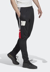 Man i svarta träningsbyxor med vita ränder, röda detaljer, nummer 10 och svarta fotbollsskor som går på en ljus bakgrund.