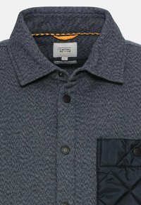 Blå quilted skjorte med et struktureret, herringbone mønster. Har sorte knapper og en ensfarvet sort lomme på venstre bryst.