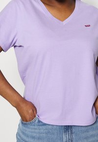 Fioletowa bawełniana koszulka z dekoltem w serek i krótkimi rękawami. Małe czerwone logo Levi's po lewej stronie, w połączeniu z jasnymi dżinsami.