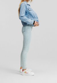 Těhotná žena v světlém modrém džínsovém bundičce, úzkých džínách a bílých teniskách s barevným vyšíváním "Karl" stojí bokem proti jednobarevnému pozadí.