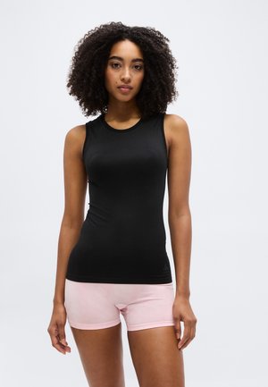 Femme aux cheveux bouclés portant un haut noir sans manches et un short rose clair, debout devant un fond gris uni.