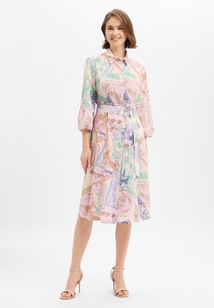 Vrouw draagt een midi-jurk met een pastelkleuren patroon, een ceintuur en driekwart mouwen, gecombineerd met beige sandalen met hoge hakken, tegen een witte achtergrond.