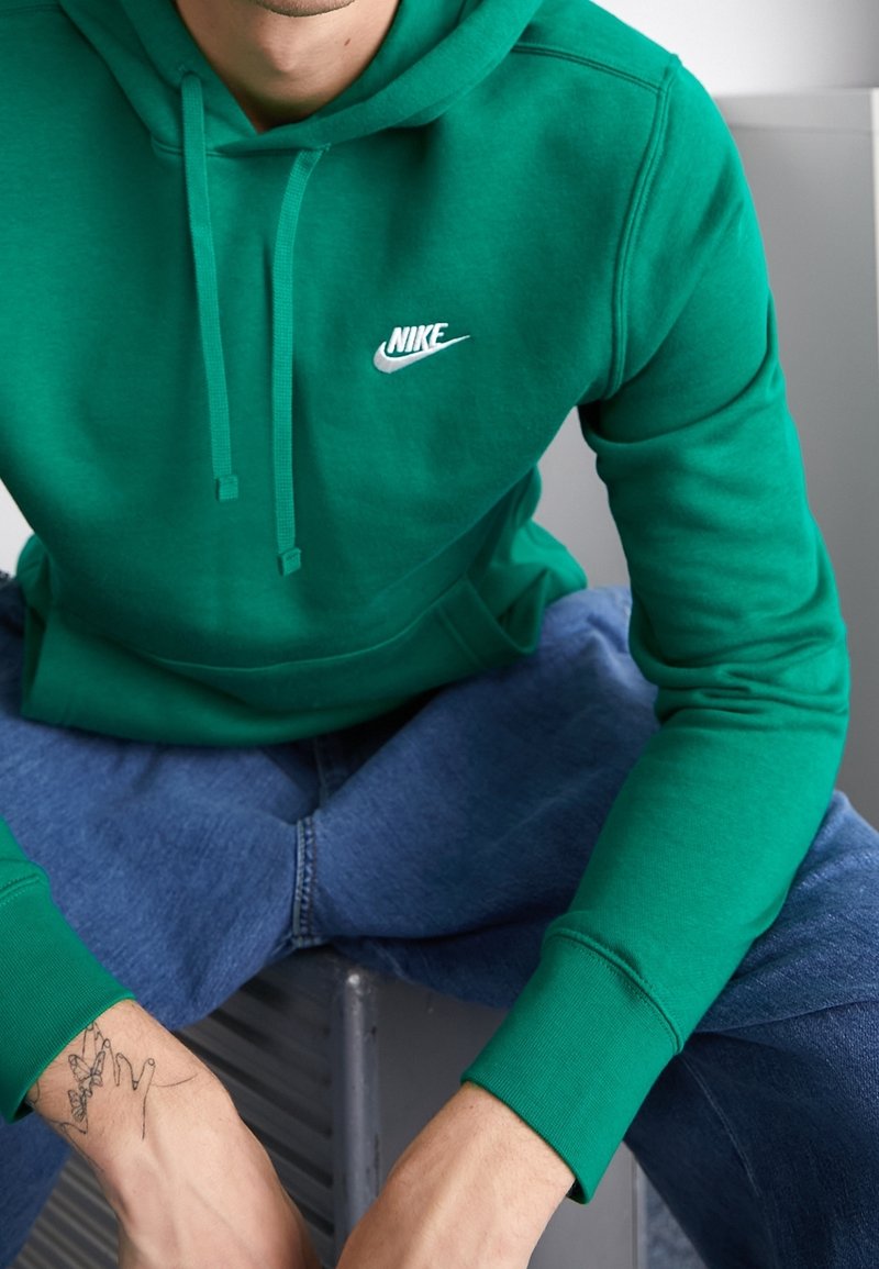 Zelená mikina Nike s kapucí, přední kapsou a stahovacími šňůrkami na kapuci, bílým logem na hrudi. Nosená s modrými džíny.