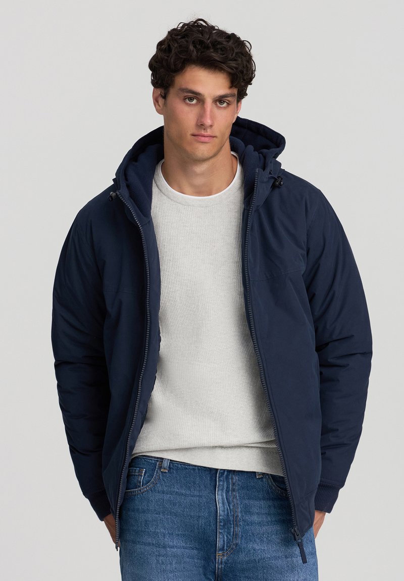 Giacca imbottita blu navy con cappuccio, chiusura zip e polsini a costine. Indossata sopra un maglione grigio chiaro a trama e jeans di denim blu.