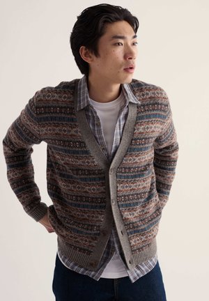 Jeune homme portant un cardigan à motifs par-dessus une chemise à carreaux et un t-shirt blanc, regardant vers la droite avec les mains derrière le dos.