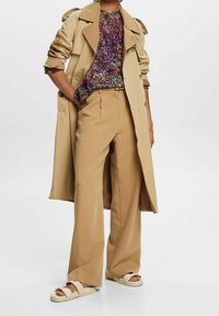 Beżowy płaszcz typu trencz na fioletowej bluzce w kwiaty, zestawiony z beżowymi szerokimi spodniami i beżowymi sandałami, podkreślając luźny, warstwowy styl.