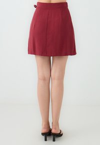 Bordeaux mini-rok met plooien, gemaakt van een lichtgewicht stof. Beschikt over een tailleband met een lusdetail. Gecombineerd met zwarte haksandalen.