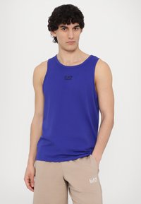 Blå ermeløs tanktopp laget av mykt stoff, har rund hals og en liten svart logo på fronten. Kombinert med lysebeige shorts.