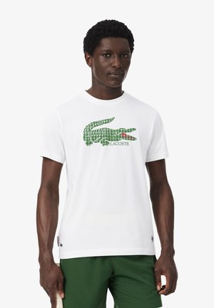 Mann trägt weißes T-Shirt mit großem grünem Krokodil-Logo und grüne Shorts, steht vor weißem Hintergrund.