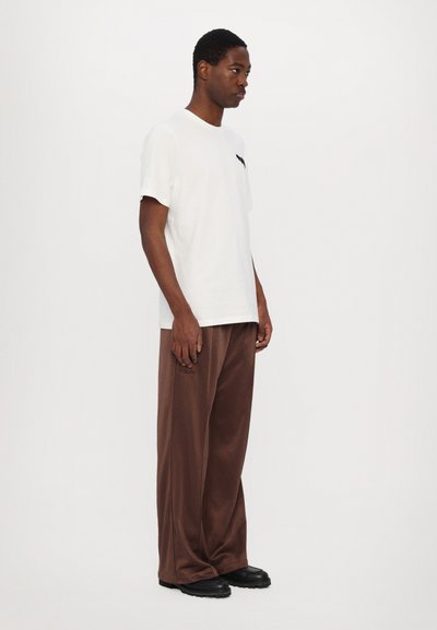 Weißes Baumwoll-T-Shirt mit kurzen Ärmeln und einem kleinen schwarzen Taschen-Detail, kombiniert mit lockeren braunen Hosen und schwarzen Schuhen.