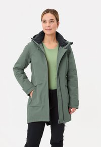 Groene waterdichte jas met een zwart gevoerde capuchon, ritszakken en een licht getailleerd ontwerp, gecombineerd met een lichtgroen topje.