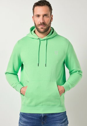 Man met baard die een lichtgroene hoodie draagt met voorzak en trekkoorden, staand tegen een effen achtergrond met zijn handen in zijn zakken.