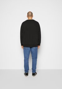 Sweat-shirt noir à manches longues, design simple. Porté avec un jean bleu et des baskets noires, vu de dos sur un fond uni.