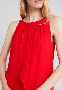 Femme portant un chemisier sans manches rouge vif avec un col froncé et un détail superposé à l'avant, sur un fond neutre.